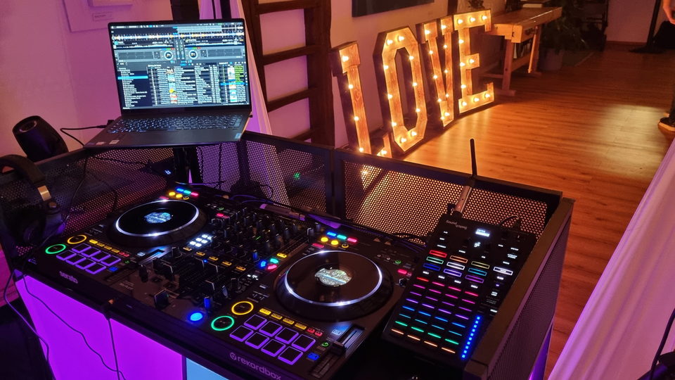 DJ-Pult mit Pioneer-Controller und Laptop, dahinter leuchtende LOVE-Buchstaben im abgedunkelten Saal