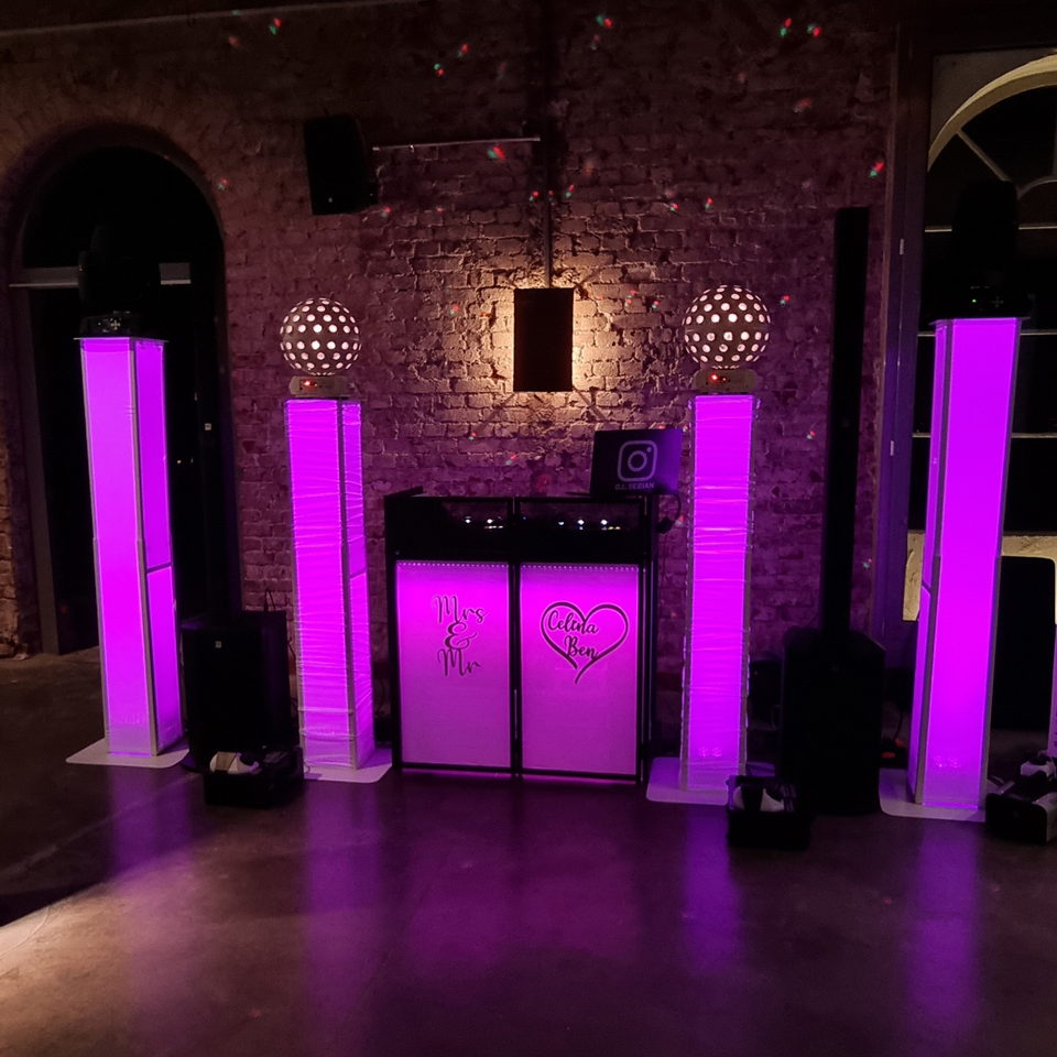 Weiß beleuchtetes DJ-Pult mit Hochzeitsmonogramm, flankiert von magentafarbenen LED-Säulen vor einer Ziegelwand