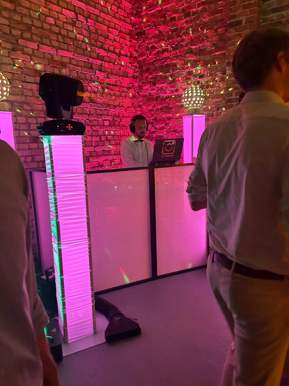 Sebastian am Pult in einem magenta ausgeleuchteten Ziegelgewölbe, davor feiernde Gäste