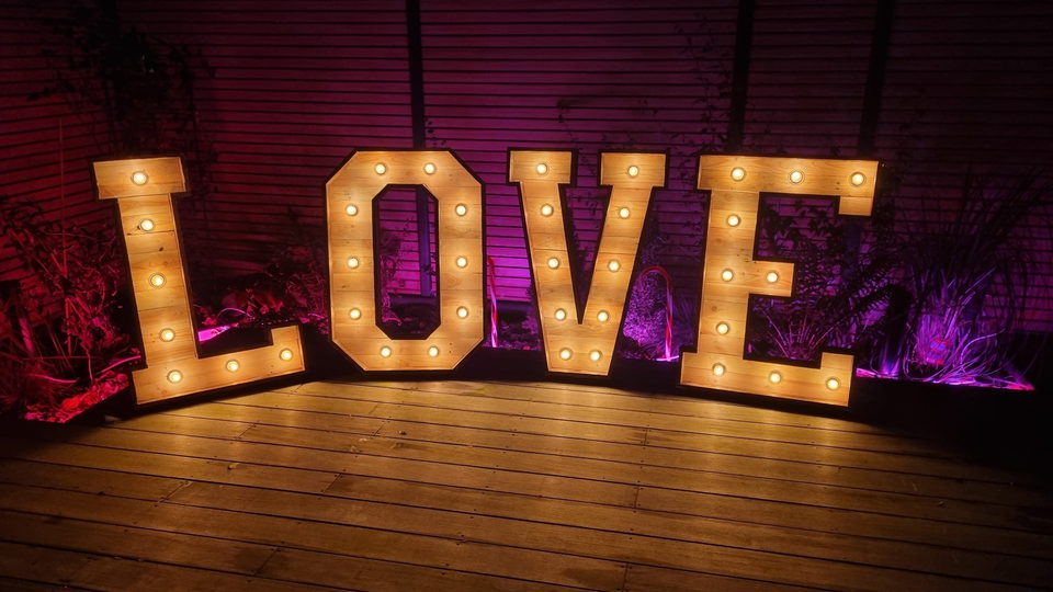 Mannshohe LOVE-Leuchtbuchstaben aus Holz mit warmen Glühbirnen auf einer Holzterrasse vor magenta angestrahlter Wand