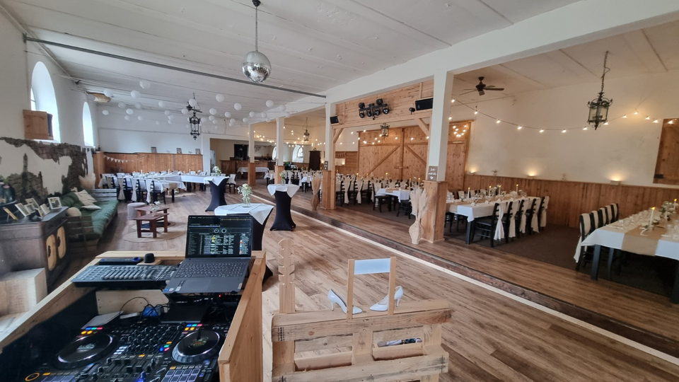 Hochzeitsscheune vor dem Fest mit eingedeckten Tafeln, Lichterketten und Discokugel, im Vordergrund das DJ-Pult